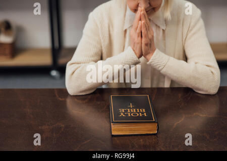 Vista ritagliata della senior donna seduta e pregando davanti della Sacra Bibbia a casa Foto Stock