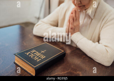 Vista parziale di senior donna seduta e pregando davanti della Sacra Bibbia a casa Foto Stock