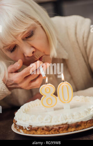 Vista ravvicinata della donna senior di illuminazione di sigaretta da candele accese sulla torta di compleanno a casa Foto Stock