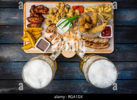 La birra snack set. Boccali di Birra e il pollo spuntini serviti sul tagliere con lo sfondo di legno. Vista superiore Foto Stock