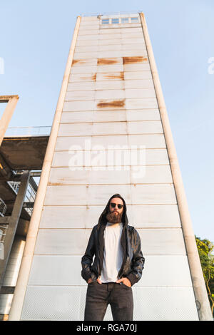 Cool barbuto giovane uomo che indossa gli occhiali da sole e giacca con cappuccio in piedi di fronte a una torre Foto Stock