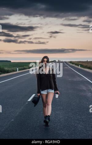 Ritratto di autostop giovane donna con zaino e bevanda camminando sulla corsia a crepuscolo serale Foto Stock