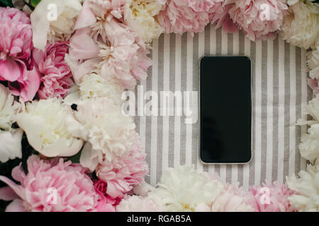 Telefono con schermo vuoto e peonie telaio laici piatto sul tavolo rustico panno. Spazio per il testo. Il blogging e instagram concetto. Biglietto di auguri mockup. Felice Foto Stock