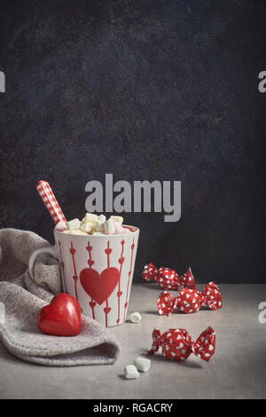 Cioccolata calda con marshmallows, cuore rosso sulla tazza, avvolto la caramella sul tavolo. Il giorno di San Valentino lo sfondo con il testo di saluto. Foto Stock