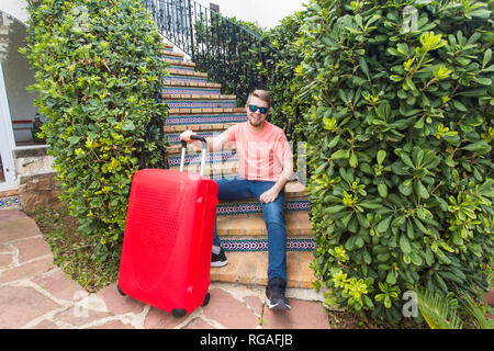 Vacanze, viaggiare e concetto di persone - uomo con red travel valigia seduti sulle scale la sua casa Foto Stock