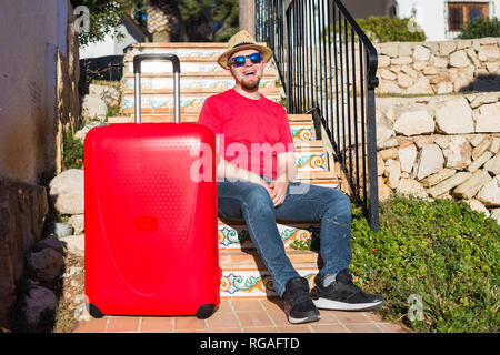 Viaggi, Turismo e concetto di persone - happy uomo seduto sulle scale in un cappello rosso con la valigia Foto Stock