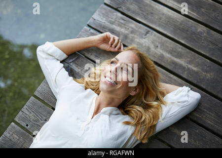 Sorridente donna bionda giacente sul molo di legno in un lago Foto Stock