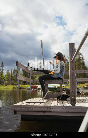 Finlandia, Lapponia, donna seduta sul pontile di un lago tramite telefono cellulare Foto Stock
