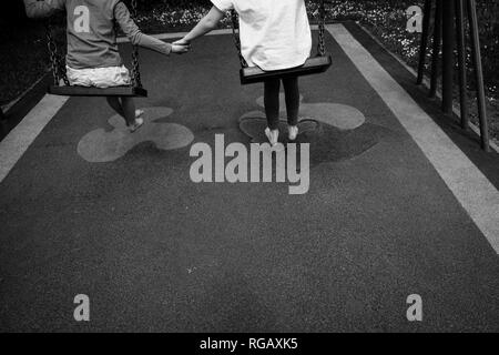 Fotografia in bianco e nero di due ragazze caucasion giocando su oscilla in un parco in un parco giochi Holding Hands Foto Stock