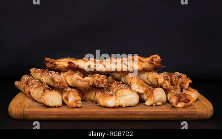 Twisted formaggio bastoncini di pane con prosciutto e i semi di sesamo su una tavola di legno su uno sfondo nero Foto Stock