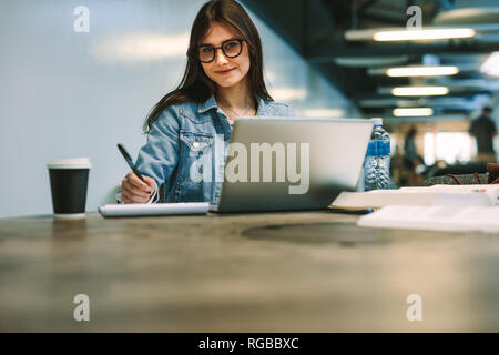 Ritratto di giovane e bella donna seduta al college biblioteca con il laptop e il blocco note. Università Femminile studente di college campus. Foto Stock