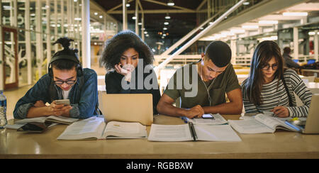 Multi-etnico gli studenti seduti al college library. Gli studenti utilizzando rendendo note, utilizzando laptop e telefoni cellulari mentre studiano insieme. Foto Stock