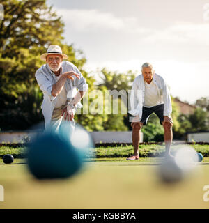 Uomo anziano giocando a bocce in un parco giochi con la sua playmate in piedi in background. Il vecchio uomo in hat gettando un Legname in blocco in un prato con boule sfocata Foto Stock