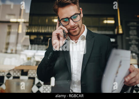 uomo d'affari seduto in un bar che passa attraverso i file dell'ufficio mentre parla al telefono cellulare. Imprenditore che gestisce il suo lavoro di lavoro seduto in un ristorante Foto Stock