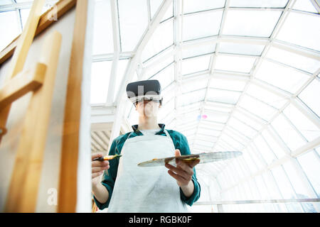 Angolo basso Ritratto di giovane uomo che indossa le cuffie VR pittura immagine sul cavalletto sotto avveniristico tetto di vetro, spazio di copia Foto Stock