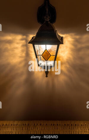 Vecchia lanterna in vetro arancione sul muro intonacato con ombre nel buio Foto Stock