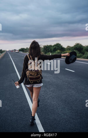 Vista posteriore del autostop giovane donna con zaino e bevanda in piedi alla linea laterale Foto Stock