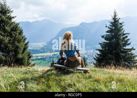 In Germania, in Baviera, Oberammergau, giovane donna escursionismo seduta sul banco di lavoro sul prato di montagna Foto Stock