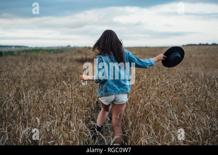 Vista posteriore della giovane donna con bevande e hat camminando nel campo di grano Foto Stock