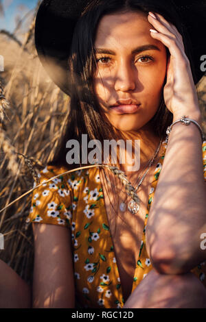 Ritratto di giovane donna che indossa abiti estivi con design floreale e un cappello seduta nel campo di grano Foto Stock