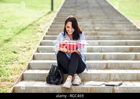 Sorridente studente seduto sulle scale all'aperto a prendere appunti in un notebook Foto Stock