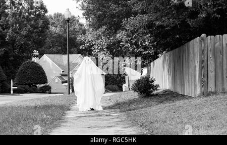 Un bianco e nero photof di un bambino in un costume fantasma realizzato a partire da un foglio di letto percorrendo a piedi il marciapiede verso la telecamera. Foto Stock