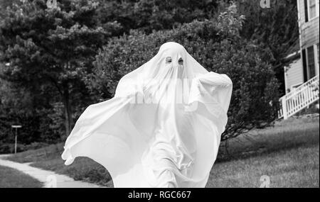 Una foto in bianco e nero di un bambino in un costume fantasma realizzate in un lenzuolo che corre lungo il marciapiede. Foto Stock