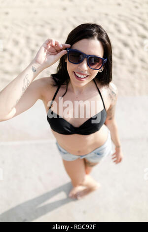 Ritratto di sorridente giovane donna con naso piercing e tatuaggio indossando occhiali da sole sulla spiaggia Foto Stock