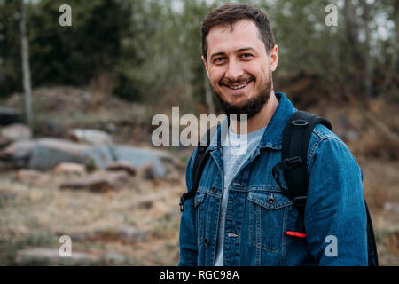 Ritratto di uomo sorridente con zaino indossando giacca denim in natura Foto Stock