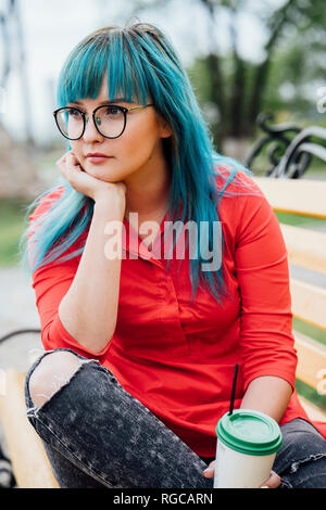 Ritratto di giovane donna con tinti di blu capelli seduta su una panchina con bevanda Foto Stock