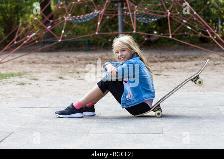 Ritratto di sorridente ragazza bionda seduta con il suo skateboard sul parco giochi Foto Stock