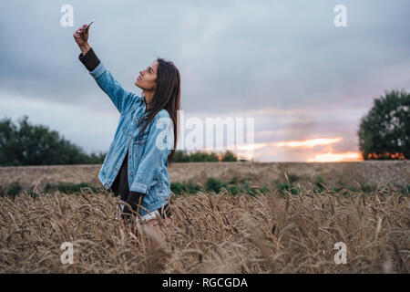 Giovane donna prendendo selfie con lo smartphone in un campo di grano al tramonto Foto Stock