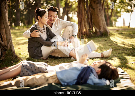 Giovani asiatici giovane seduto sull'erba nel parco chiacchierando con due bambini che giace la lettura di libro in primo piano. Foto Stock