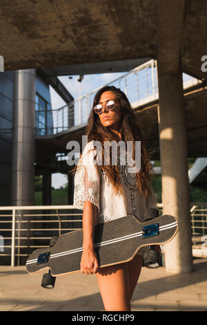 Giovane donna in piedi con lo skateboard in città Foto Stock