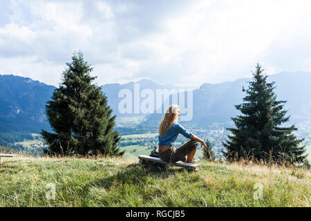 In Germania, in Baviera, Oberammergau, giovane donna escursionismo seduta sul banco di lavoro sul prato di montagna Foto Stock