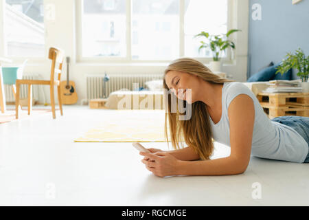Sorridente giovane donna sdraiata sul pavimento nel suo loft guardando al telefono cellulare Foto Stock