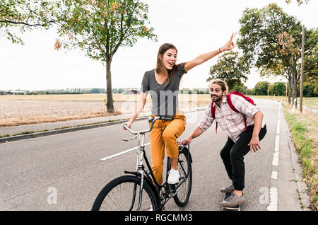 Felice coppia giovane con noleggio biciclette e skateboard sulla strada di campagna Foto Stock