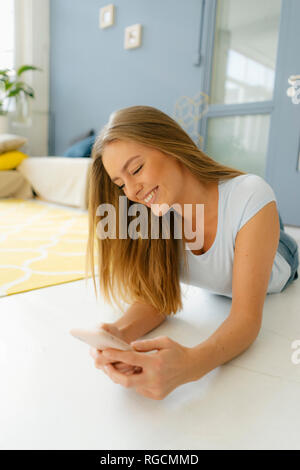 Sorridente giovane donna sdraiata sul pavimento nel suo loft guardando al telefono cellulare Foto Stock