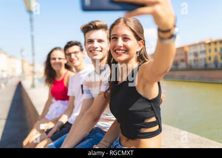 L'Italia, Pisa, gruppo di quattro amici felice seduto su una parete lungo il fiume Arno prendendo un selfie Foto Stock