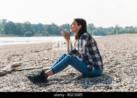 Rilassata giovane donna tenendo un mug seduto in riva al fiume Foto Stock