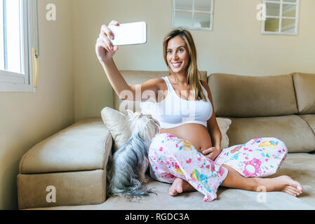 Sorridente donna incinta prendendo un selfie con il cane sul lettino a casa Foto Stock