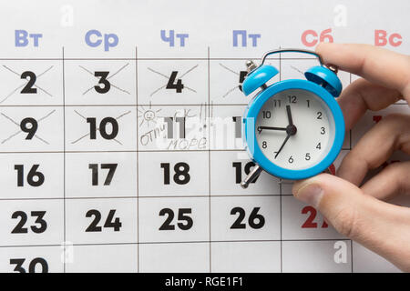 Una mano trattiene una piccola sveglia, in background è un calendario con il giorno segnato e la parola vacanza Foto Stock
