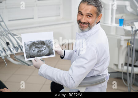 Allegro dentista sorridente, che pongono e che mostra apparecchiature odontoiatriche a raggi x della ganascia. Stomatologist seduto in poltrona guardando la fotocamera. Specialista professionale che indossa in bianco uniforme mentre si lavora in clinica. Foto Stock