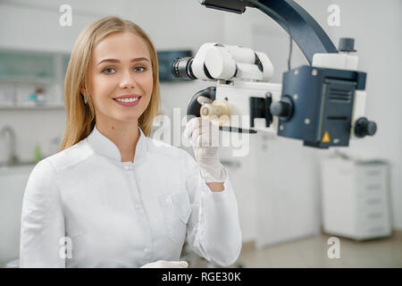 Vista frontale della bellissima femmina dentista in bianco uniforme guardando la fotocamera e sorridente. Giovane medico seduto a lavorare in studio dentistico e mantenendo le attrezzature moderne. Concetto di stomatologia. Foto Stock