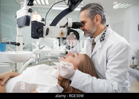 Dentista maschio mediante microscopio dentale per denti esame. Bella donna sdraiata con la bocca aperta sulla poltrona del dentista. Maturo uomo barbuto che indossa in bianco cappotto medico. Foto Stock