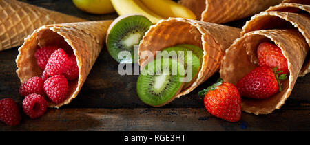 Frutta fresca tropicale visualizzati nella corni o waffle coni gelato riversandosi su legno rustico con kiwi, fragole lamponi di annunci Foto Stock