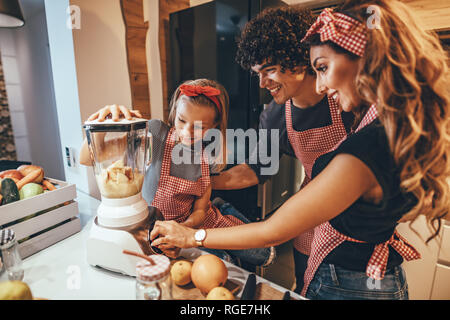 Felice famiglia giovane è la preparazione di pasto sano in cucina. Madre, padre e figlia stanno mettendo fette di frutta nel frullatore e messa a frutto s Foto Stock