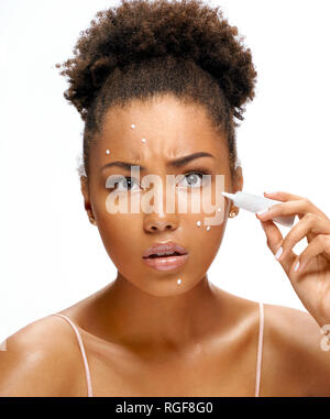 Scowling ragazza l'applicazione di crema di trattamento. Foto di African American Girl su sfondo bianco. La cura della pelle e concetto di bellezza Foto Stock