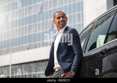 Giovane americano africano uomo in piedi vicino a car mani nelle tasche in cerca riposo sorridente pensieroso vista laterale Foto Stock