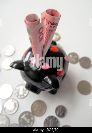 Nero salvadanaio riempito con il cinese 100 RMB banconote e sparse monete cinesi su un bianco sullo sfondo di legno. Il 2019 è l'anno del maiale. Foto Stock